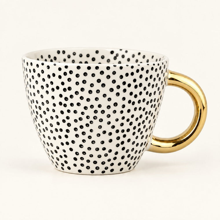 A white ceramic coffee mug covered in small black polka dots, featuring a shiny gold handle, set against a plain