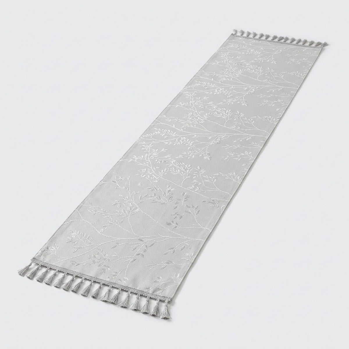 A long, narrow white fabric runner with a subtle tonal floral pattern and tassel fringe on both ends, shown on a light