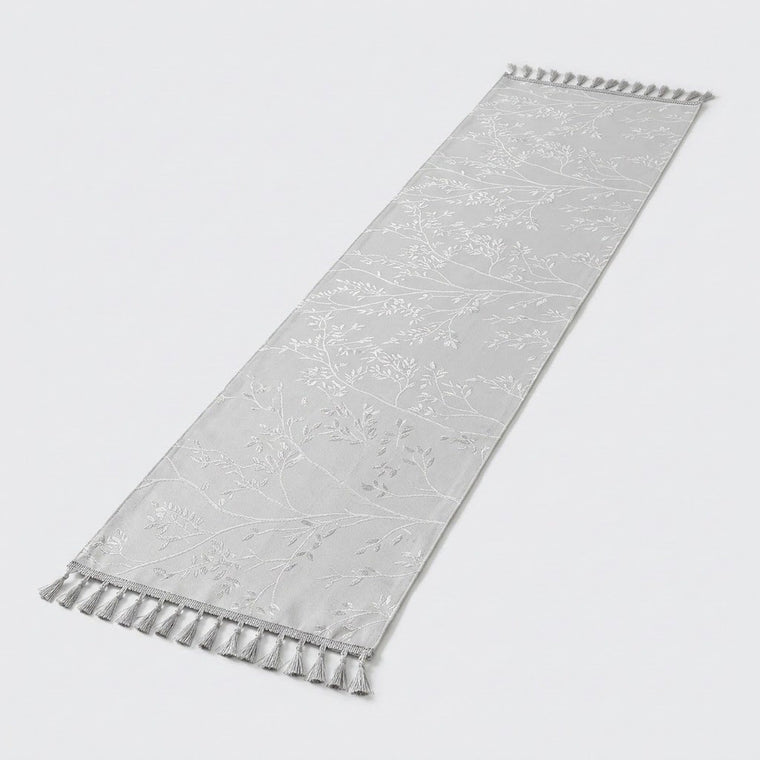 A long, narrow white fabric runner with a subtle tonal floral pattern and tassel fringe on both ends, shown on a light