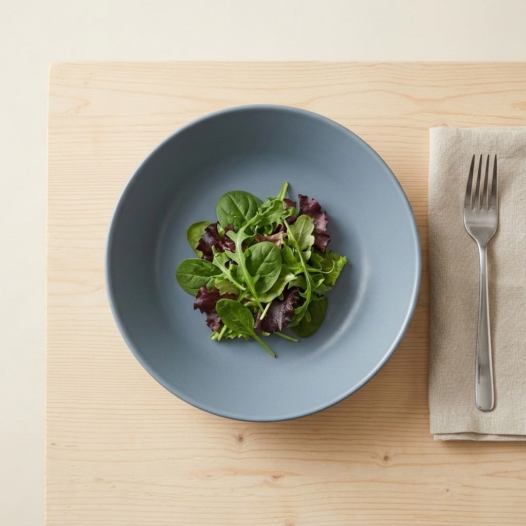 A matte slate blue bowl filled with a green and purple salad, placed on a light wood placemat next to a folded beige