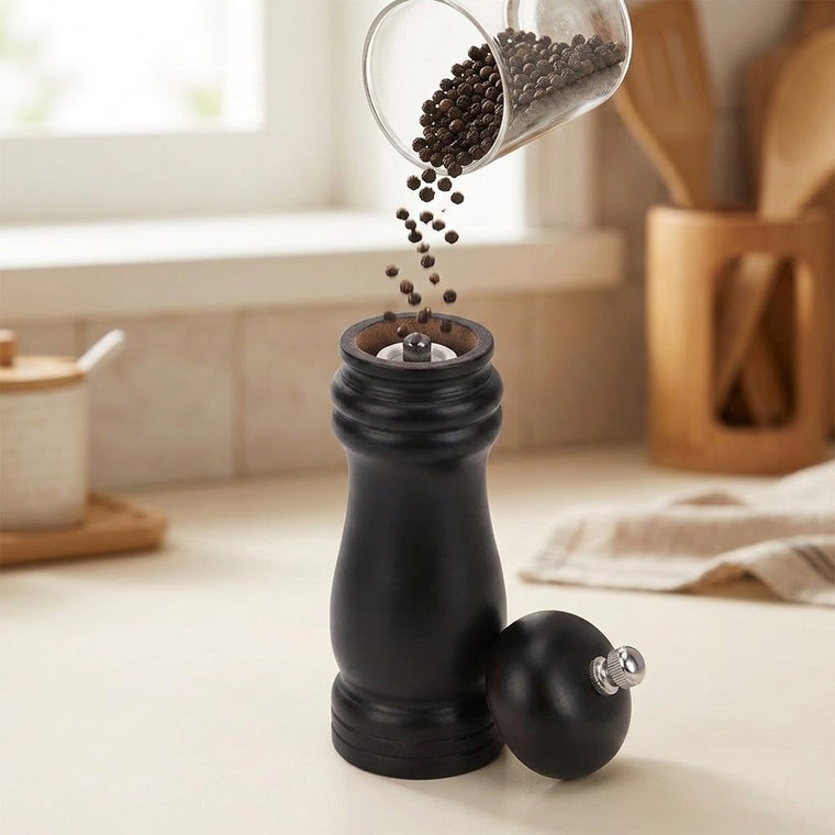 A black pepper mill grinder with a matching round ball and a clear container pouring peppercorns into the open top, shown