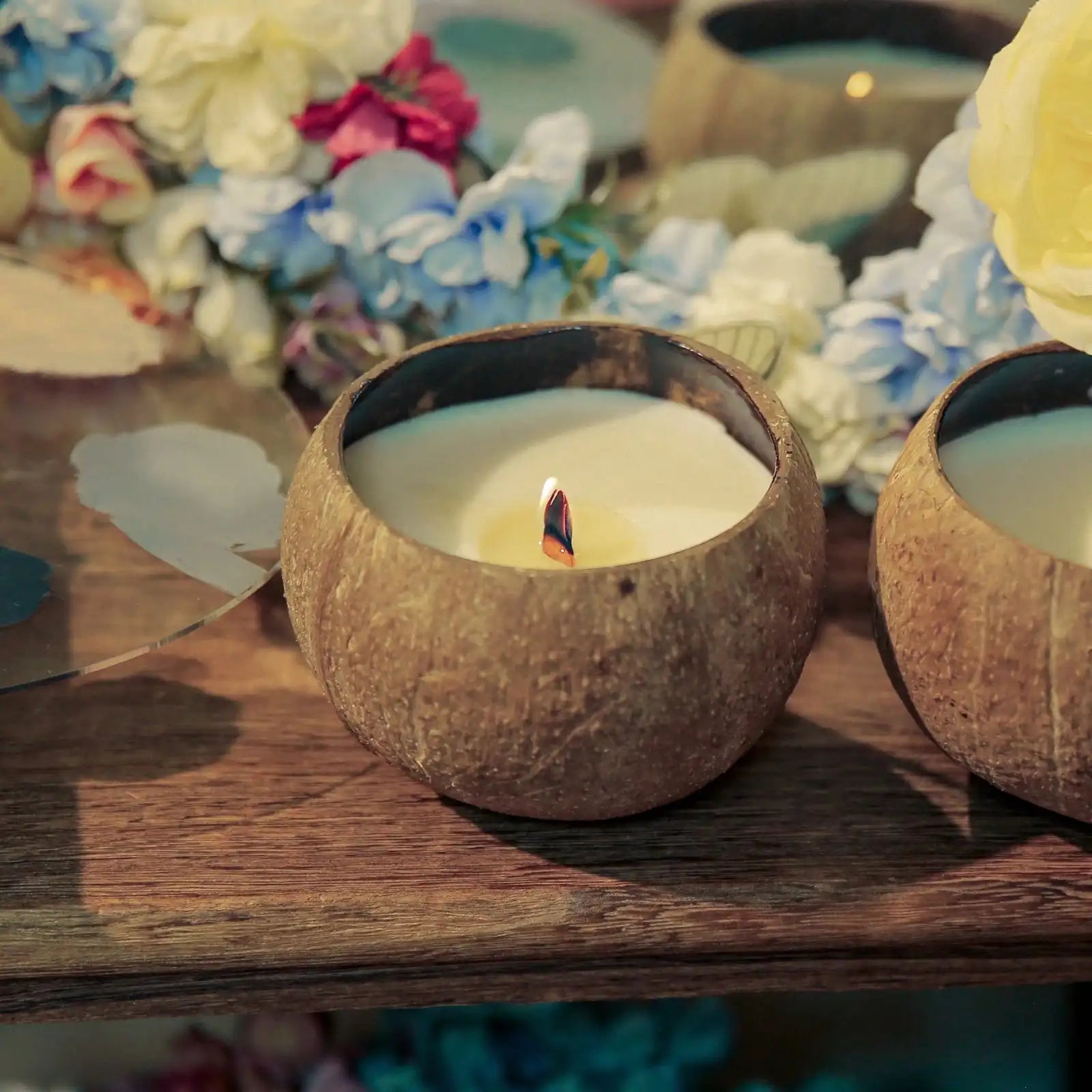 Whole coconut shell candle burning on a wooden table with flowers behind