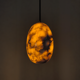 An oval pendant light with a glossy, marbled amber and black resin finish, suspended by a thin black cord against a dark, neutral background.