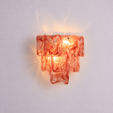 A modern tiered wall sconce light fixture made of textured, translucent orange-red glass, glowing with a warm light against a plain light gray background.