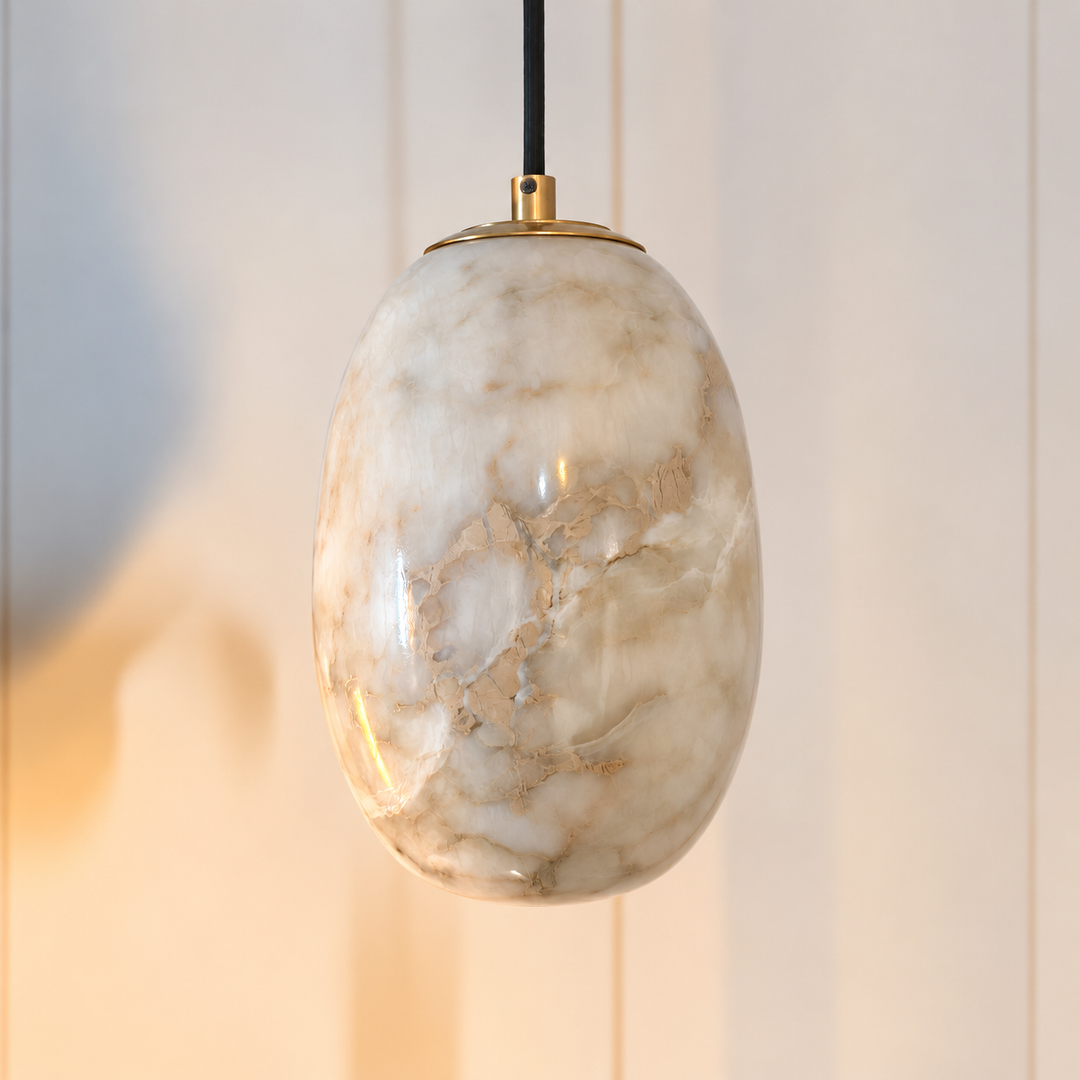 An oval pendant light with a polished white and beige marble glass shade, suspended by a black cord with a gold top cap, set against a softly blurred light beige paneled background.