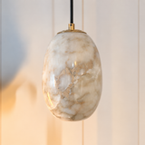An oval pendant light with a polished white and beige marble glass shade, suspended by a black cord with a gold top cap, set against a softly blurred light beige paneled background.