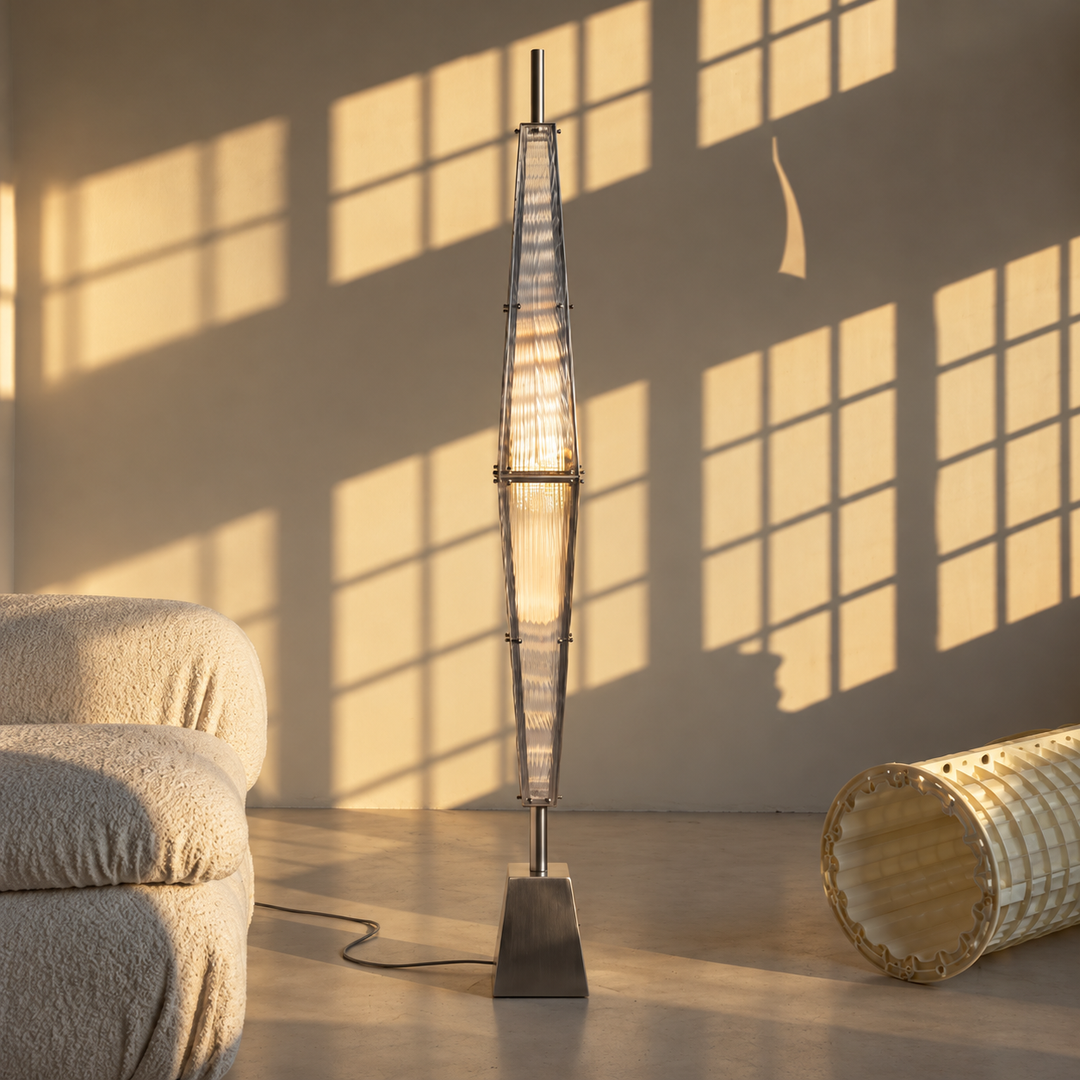 A tall, modern floor lamp with a cylindrical glass shade and a square base, standing in a minimalist room with warm sunlight casting windowpane shadows on the wall and floor.