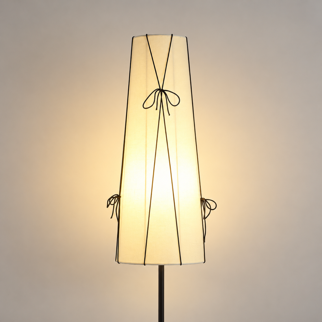 A modern floor lamp with a tapered, pleated off-white fabric shade decorated with thin black cord bows, shown illuminated against a warm neutral background.