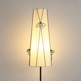 A modern floor lamp with a tapered, pleated off-white fabric shade decorated with thin black cord bows, shown illuminated against a warm neutral background.