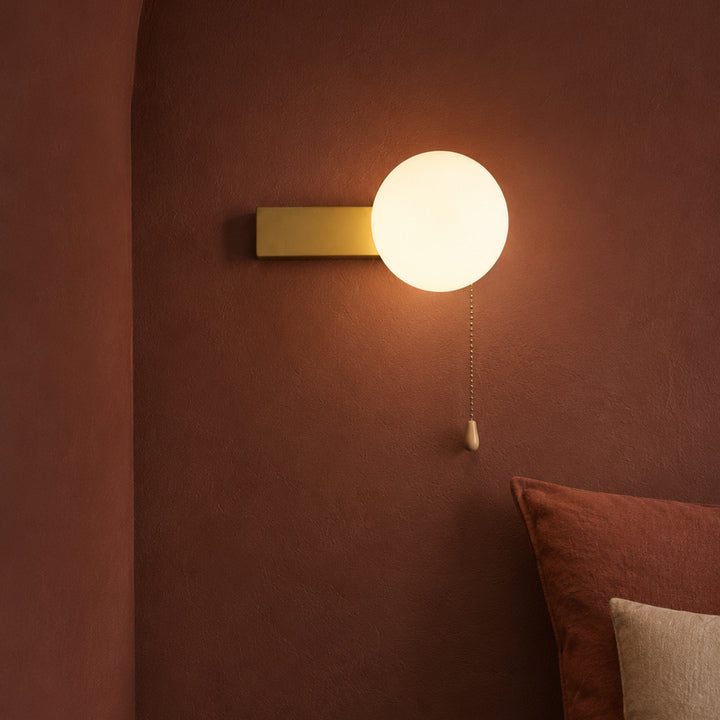 A modern wall-mounted light fixture with a glowing spherical frosted globe and a rectangular gold backplate, mounted on a textured terracotta wall above a bed.