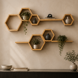 Hexagonal wooden floating wall shelves mounted on a beige wall above a console table, styled with small potted plants and ceramic vases, with a beige sofa and dark pillows in the foreground.