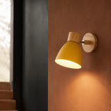 A modern wall sconce light fixture with a matte yellow shade and natural wood accents, mounted on a textured terracotta wall and casting a warm glow.
