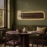 A modern wall-mounted wooden shelf with integrated warm LED backlighting, mounted on an olive green wall above a matching upholstered banquette in a dining space.