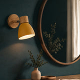 A modern wall sconce with a warm yellow dome shade and natural wood mounting plate and neck, shown lit on a dark teal wall next to a large round wooden mirror.