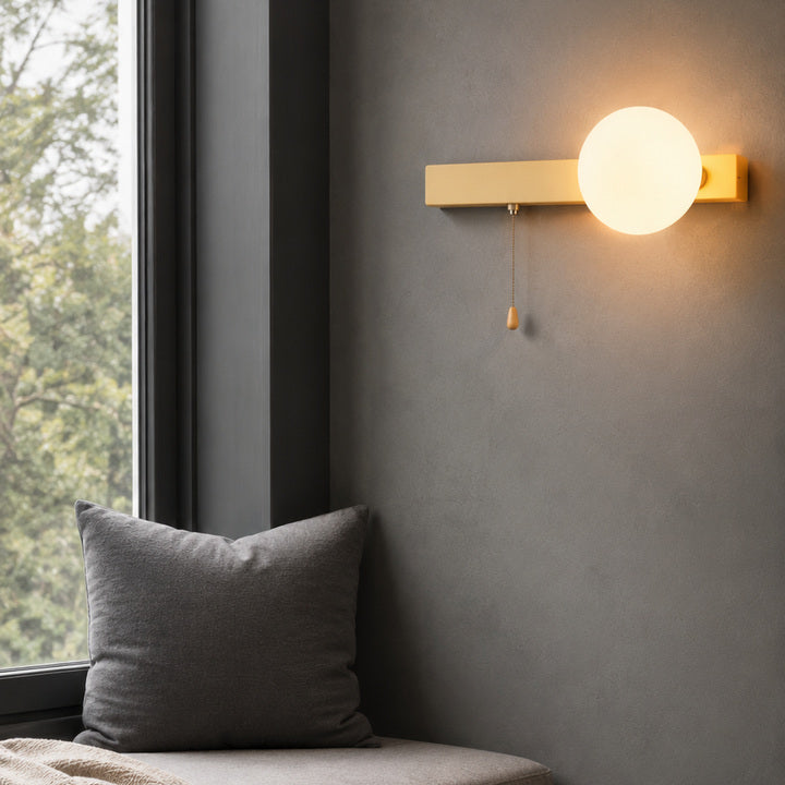 A modern wall-mounted light fixture with a glowing frosted globe and a pull chain, mounted on a textured gray wall in a styled interior setting with a window and a gray pillow.