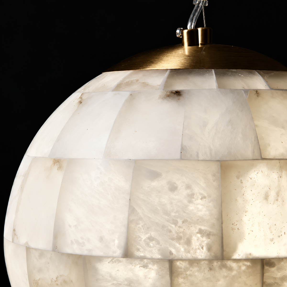 Close-up of a spherical pendant light shade made of stacked translucent white stone tiles, with a warm brass metal cap and hanging cord visible against a black background.