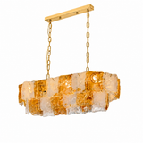 A linear chandelier with layered amber and clear textured glass hangs from gold chains against a white background.