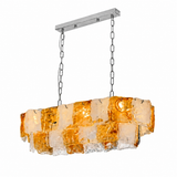A linear chandelier with layered amber and clear textured glass hangs from silver chains against a white background.