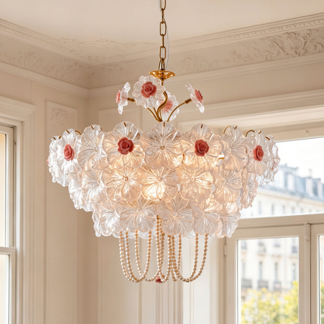 A vintage-style floral chandelier with clear glass flower petals, pink rose centers, and gold-toned metal hardware, hanging in a classic cream-colored room with ornate molding and windows.