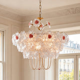 A vintage-style floral chandelier with clear glass flower petals, pink rose centers, and gold-toned metal hardware, hanging in a classic cream-colored room with ornate molding and windows.