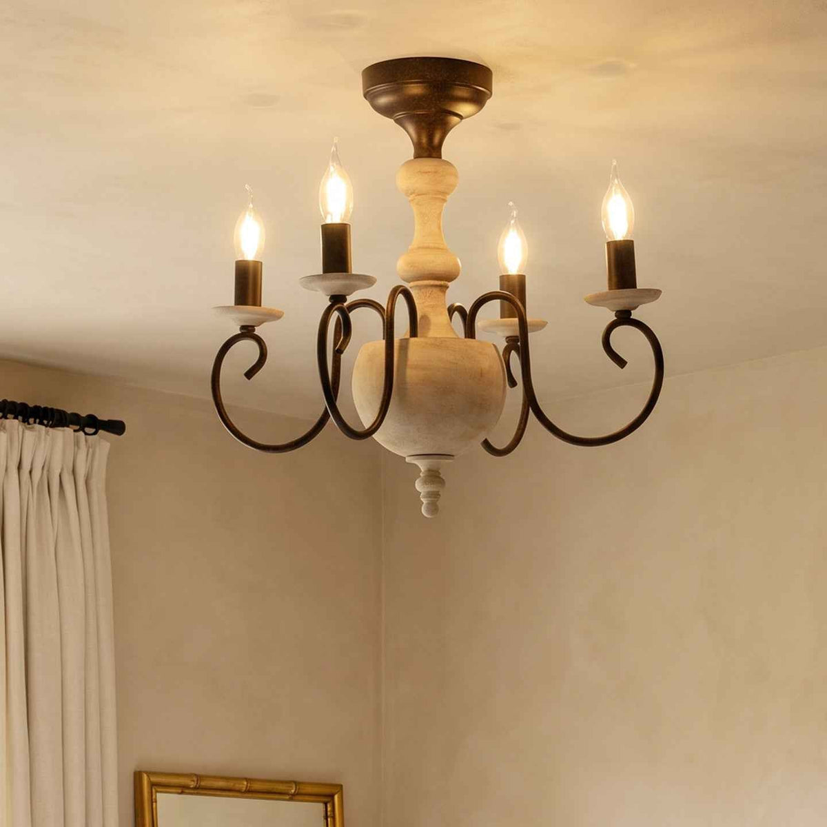 Four-light candle chandelier with a distressed cream wooden body and dark iron arms hanging near cream curtains in a warm room.