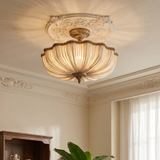 A semi-flush mount ceiling light fixture with a pleated glass shade and ornate brass fixture, mounted on a decorated cream-colored ceiling in a classic room setting.