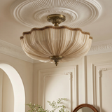 A vintage-style semi-flush mount ceiling light fixture with a pleated cream shade and antique brass hardware, shown installed on a decorative white plaster ceiling in an elegant room.