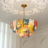 A modern chandelier with a gold-toned brass canopy and chain, featuring multiple textured, translucent glass shades in gold, blue, red, and white, hanging in an elegant room with a white ornate plaster ceiling.