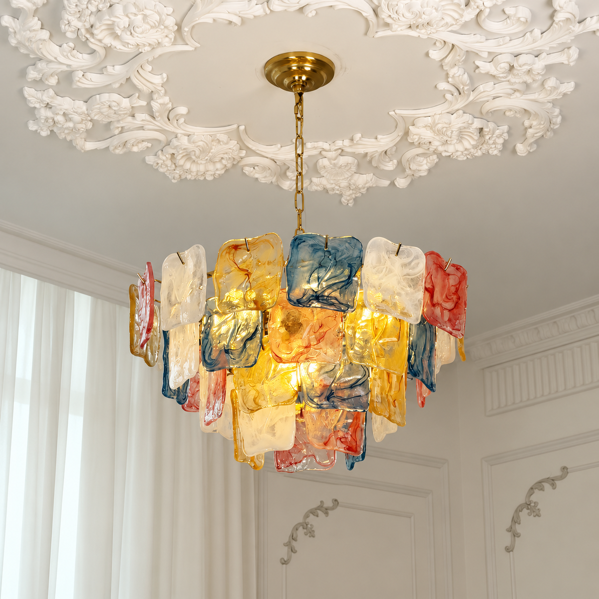 A modern chandelier with a gold-toned brass canopy and chain, featuring multiple textured, translucent glass shades in gold, blue, red, and white, hanging in an elegant room with a white ornate plaster ceiling.