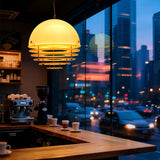 A modern pendant light with a warm glowing dome shade and horizontal slat details hangs over a wooden coffee bar inside a cafe, with a blurred city street scene visible in the background window.