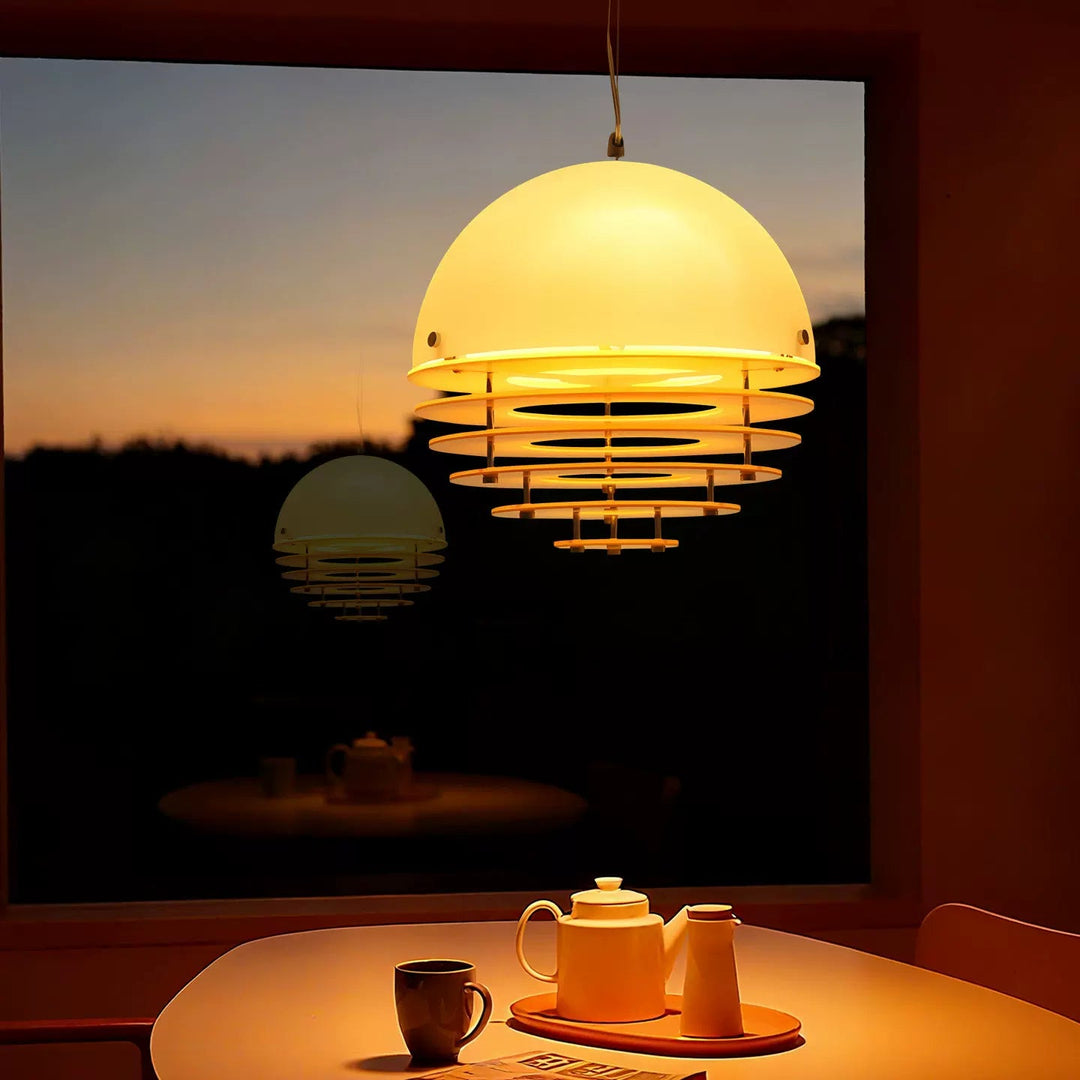 A modern pendant light with a glowing warm white dome shade and horizontal metal rings, hanging over a table with a teapot and cup in a dimly lit room at dusk.