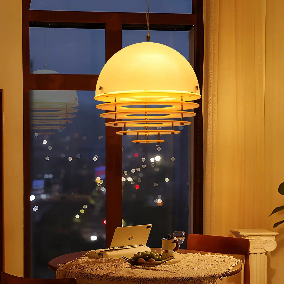A modern dome-shaped pendant light with a glowing warm white shade and horizontal metallic rings, hanging over a dining table in a warmly lit room with a large window showing a city night view.