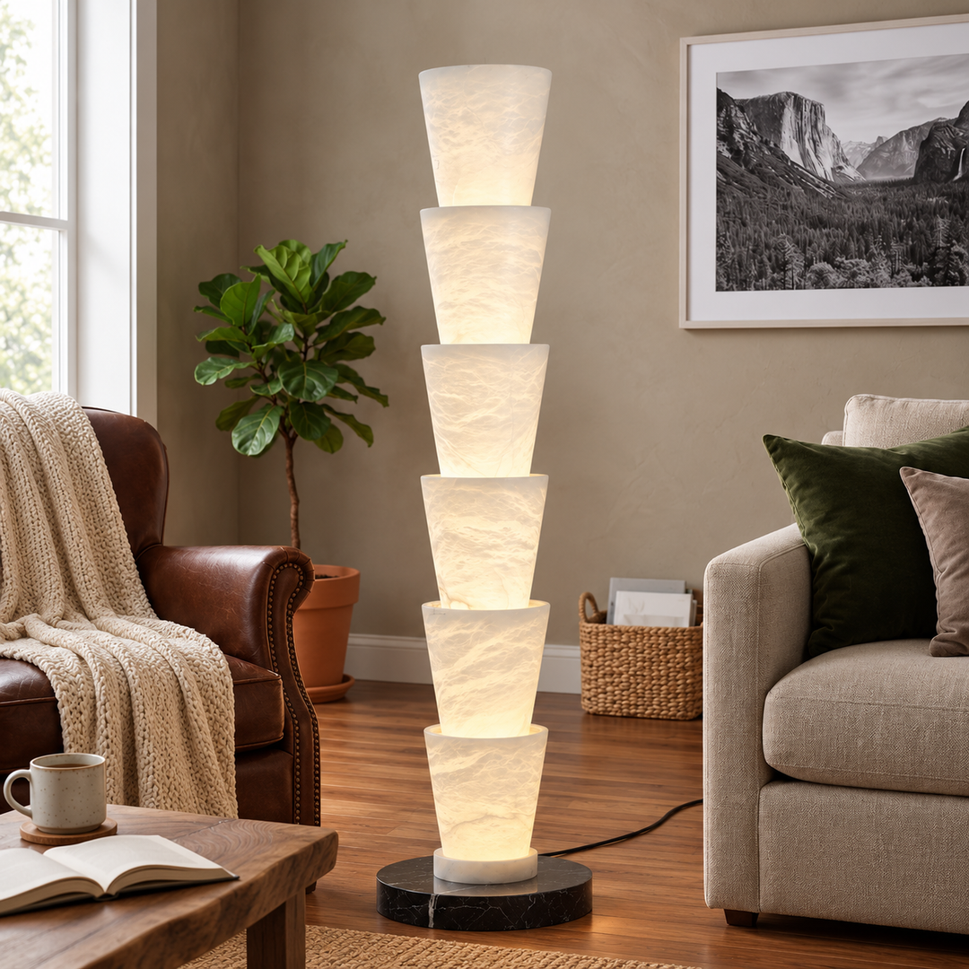 A modern spiral floor lamp with seven stacked, tapered white textured shades glowing from within, set on a black round base in a cozy living room next to a brown leather armchair and a beige sofa.