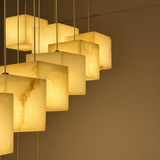 Chandelier close-up showing alabaster cube shades glowing warm yellow on thin suspension cables against a dark background.