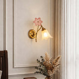 A wall-mounted sconce with a gold brass finish, featuring a pink flower-shaped top and a glowing bell-shaped glass shade, installed on a beige paneled wall next to a window.