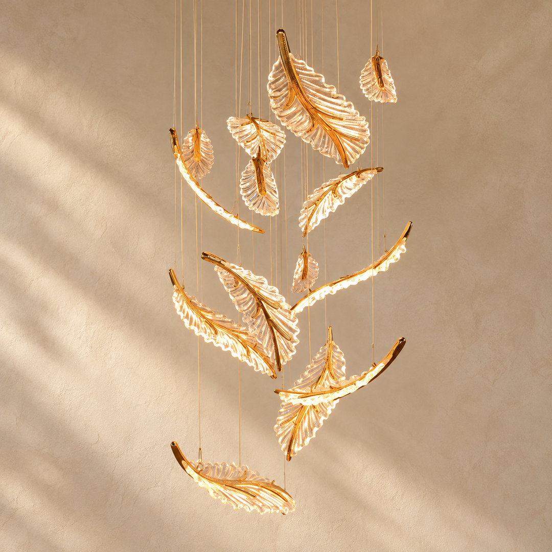 A modern hanging chandelier featuring multiple translucent leaf-shaped glass shades with warm gold-toned metal edging, suspended from thin cables against a textured beige wall with soft shadow patterns.