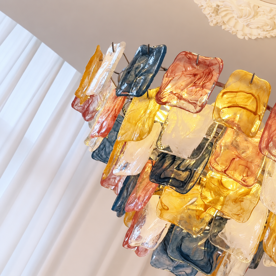 A modern chandelier featuring multiple rectangular glass panels in amber yellow, dusty pink, slate blue, and translucent white, suspended by thin wires against a white interior wall.