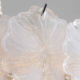 Close-up of a decorative chandelier light with translucent white glass petals and subtle gold glitter accents, showing a hanging metal rod against a soft gray background.