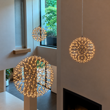 Three spherical geometric pendant lights with a gold-toned metal frame and warm white bulbs, shown hanging in a modern interior with beige walls, windows, and a fireplace.