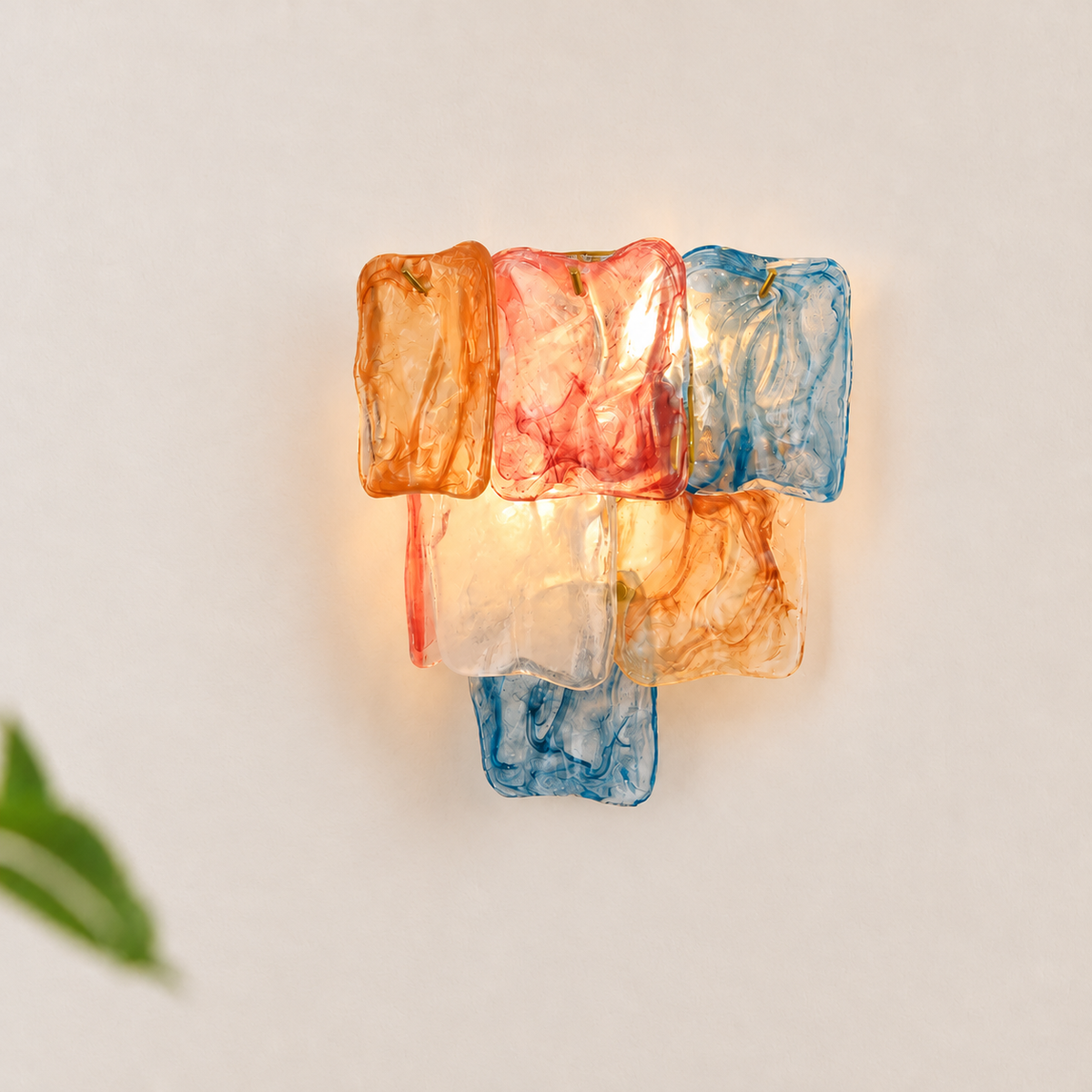 Wall sconce with orange, pink, blue, and clear glass panels glowing against a pale wall near green leaves.