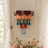 A multicolored ceramic and glass wall sconce mounted on a cream paneled wall between lace curtains, above pink roses.