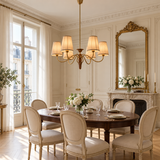 A luxury dining room featuring a round dark wood dining table surrounded by eight upholstered cream chairs, under a multi-light chandelier. The room has classic cream walls with molding, a large gold-framed mirror, and a fireplace with a floral arrangement.