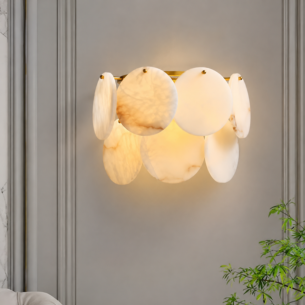 Illuminated alabaster and brass wall sconce on a gray paneled wall beside a leafy plant.