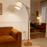 A modern floor lamp with a warm wood-look frame and a white drum shade stands in a living room next to a brown velvet sofa, casting a soft glow in a styled interior.
