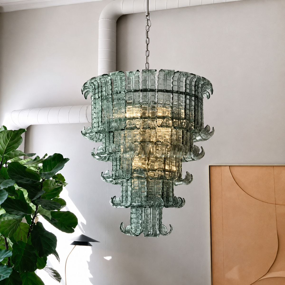 A tiered glass chandelier made of translucent green glass, hanging in a room with white walls, exposed ductwork, a green plant, and a tan abstract wall art piece.