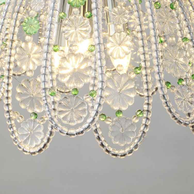 A close-up of a clear crystal chandelier shade featuring intricate floral cutouts, hanging beaded strands, and small green faceted crystal accents against a neutral gray background.
