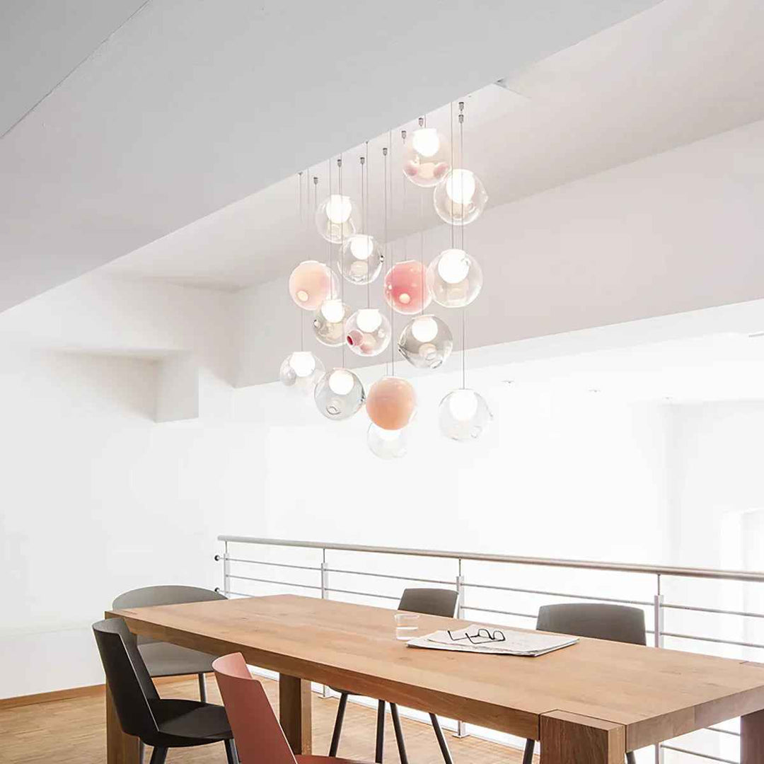 A modern multi-light chandelier with hanging clear glass globes and soft pink and peach illuminated bulbs, suspended over a wooden dining table in a bright minimalist interior.