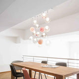 A modern multi-light chandelier with hanging clear glass globes and soft pink and peach illuminated bulbs, suspended over a wooden dining table in a bright minimalist interior.