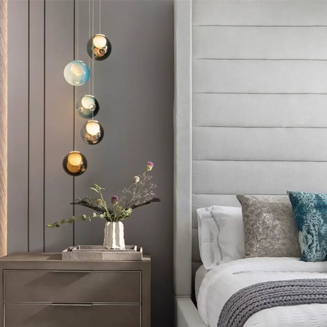 A chandelier with five tinted glass globes hangs beside a bed above a nightstand holding a vase of flowers.