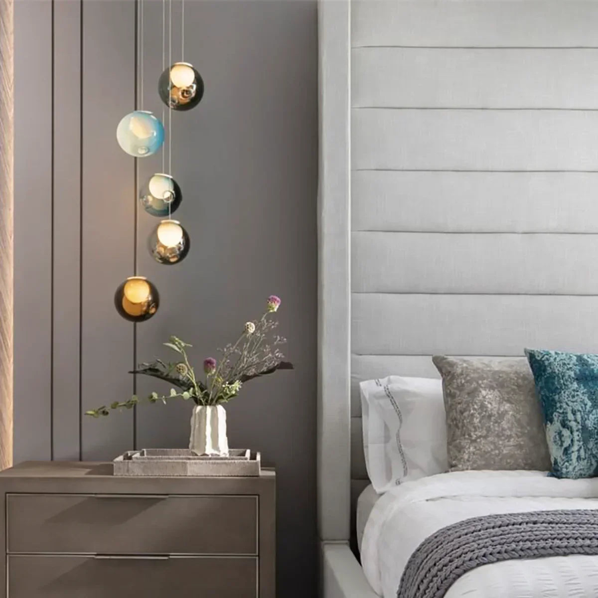 A chandelier with five tinted glass globes hangs beside a bed above a nightstand holding a vase of flowers.